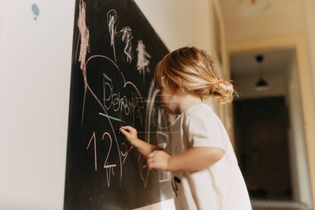 Téléchargez les photos : Une petite fille dessinant sur un tableau à la craie. L'enfant créatif joue à la maison, apprenant à dessiner des formes de base et des premières lettres. - en image libre de droit