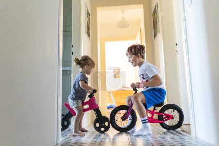 Foto de Los hermanos están montando bicicletas en el interior, en el pasillo de la casa. Un niño y una niña se están divirtiendo durante las vacaciones en casa, compitiendo entre sí en bicicletas. Hermano y hermana sentados en bicicletas - Imagen libre de derechos