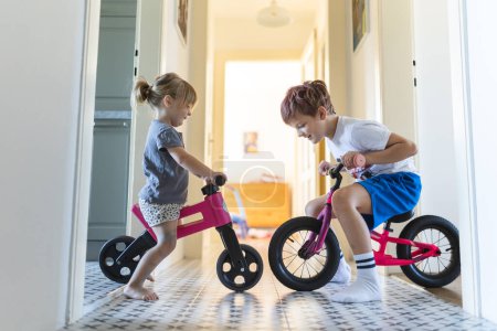 Foto de Los hermanos están montando bicicletas en el interior, en el pasillo de la casa. Un niño y una niña se están divirtiendo durante las vacaciones en casa, compitiendo entre sí en bicicletas. Hermano y hermana sentados en bicicletas - Imagen libre de derechos