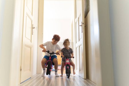 Téléchargez les photos : Les frères et sœurs font du vélo à l'intérieur, dans le couloir de la maison. Un jeune garçon et une jeune fille s'amusent pendant les vacances à la maison, se faisant la course à bicyclette. Concept d'amour fraternel et fratrie - en image libre de droit