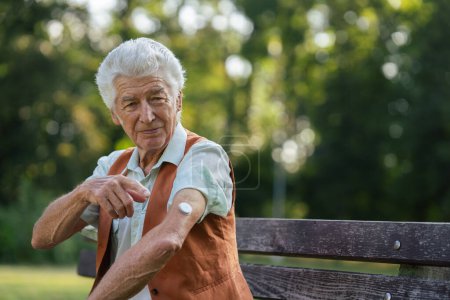 Téléchargez les photos : Un senior montrant un détecteur de glucose continu sur son bras. Homme diabétique vérifiant sa glycémie à l'extérieur, dans le parc de la ville. - en image libre de droit