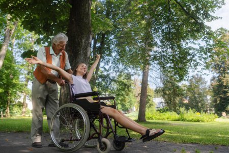 Photo for The senior man pushing young caregiver in a wheelchair, having fun. The elderly man feeling good and fit, he is able to push his granddaughter in the wheelchair. - Royalty Free Image