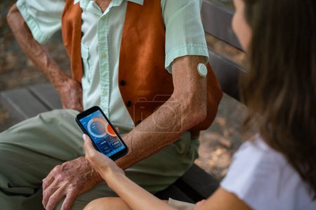 Aide soignant homme diabétique aîné vérifier ses données de glucose sur smartphone. Aîné diabétique utilisant un glucomètre continu. Petite-fille enseignant à son grand-père à utiliser un smartphone, tapant des textes