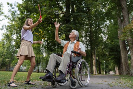 Foto de El hombre en silla de ruedas divirtiéndose con su cuidador en el parque, atrapando burbujas. El anciano en silla de ruedas realiza ejercicios sencillos, moviéndose mientras atrapa burbujas sopladas por la joven enfermera. - Imagen libre de derechos