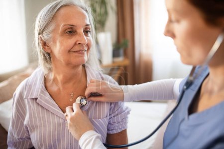 Close-up of nurse examining senior patient with stethoscope at home. Female doctor listening breathing and heart of elderly patient. Check-up after heart surgery.