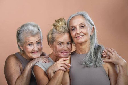 Foto de Retrato de estudio de tres hermosas mujeres mayores con pelos grises. Mujeres ancianas amigas abrazándose mirando a la cámara. Aislado sobre un fondo marrón rosado. Copiar espacio. - Imagen libre de derechos
