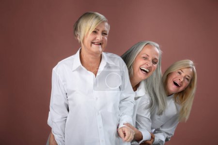 Foto de Retrato de estudio de tres hermosas mujeres mayores con pelo gris en camisa blanca para hombre. Ancianos amigos divertidos parados uno detrás del otro, riendo. Aislado sobre un fondo marrón rosado. Copiar espacio. - Imagen libre de derechos