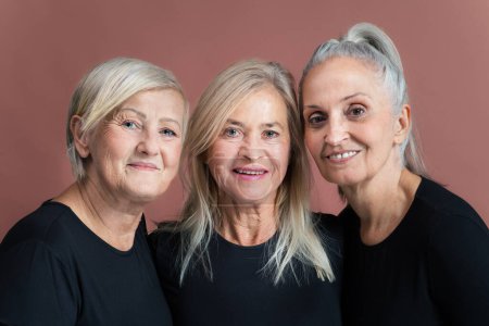 Téléchargez les photos : Portrait studio de trois belles femmes âgées aux cheveux gris en hauts noirs. Des amies âgées se tenant l'une l'autre regardant la caméra. Isolé sur un fond rose brun. Espace de copie. - en image libre de droit