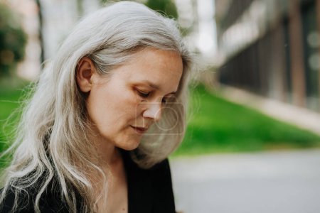 Téléchargez les photos : Portrait de femme mûre coûteuse d'âge moyen avec de longs cheveux gris, debout en plein air dans la ville. - en image libre de droit