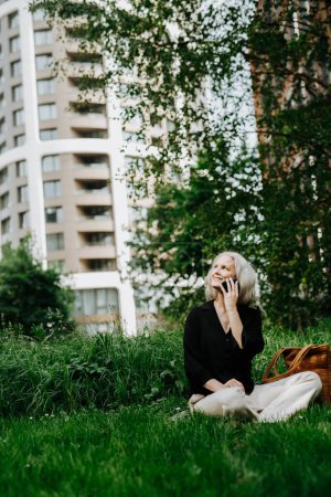 Téléchargez les photos : Portrait de belle femme mûre d'âge moyen avec de longs cheveux gris, assise sur l'herbe dans le parc de la ville. Femme souriante assise les jambes croisées sur l'herbe, faisant un appel. - en image libre de droit
