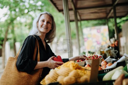 Téléchargez les photos : Portrait d'une belle femme mûre faisant du shopping au marché de la ville. Femme d'âge moyen achetant des légumes et des fruits frais au stand du marché. - en image libre de droit