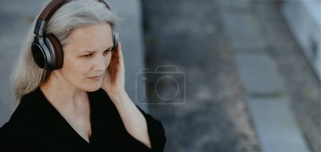 Téléchargez les photos : Plan de tête d'une belle femme aux cheveux gris, assise sur des escaliers en béton dans la ville, écoutant de la musique à travers des écouteurs sans fil. Bannière avec espace de copie. - en image libre de droit