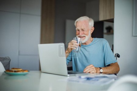 Homme âgé en fauteuil roulant travaillant à domicile pendant sa retraite. Homme âgé utilisant les technologies numériques, travaillant sur un ordinateur portable, filmant quelqu'un. Concept de seniors et compétences numériques.