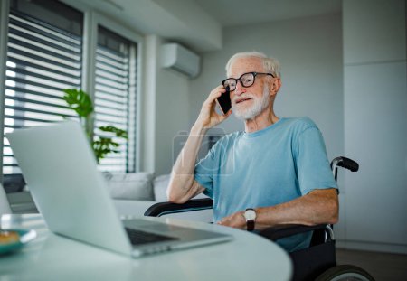 Senior man in a wheelchair working from home during retirement. Elderly man using digital technologies, working on a laptop and making phone call at home. Concept of seniors and digital skills.