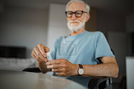 Téléchargez les photos : Homme âgé en fauteuil roulant prenant des pilules à la maison. Homme chroniquement malade prenant des médicaments, lisant l'étiquette sur le flacon. - en image libre de droit