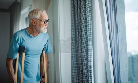 Téléchargez les photos : Triste homme âgé avec des béquilles passer du temps seul dans son appartement, regardant par la fenêtre. Concept de solitude et de dépendance des retraités. - en image libre de droit