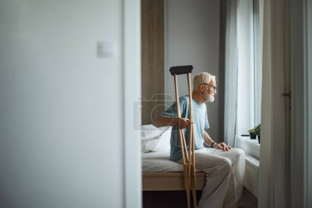Téléchargez les photos : Triste homme âgé avec des béquilles passer du temps seul dans son appartement, regardant par la fenêtre. Concept de solitude et de dépendance des retraités. - en image libre de droit