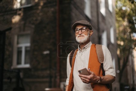 Téléchargez les photos : Touriste senior explorer une nouvelle ville et visiter des sites intéressants. Homme âgé se promenant dans la ville tout en tenant un smartphone. - en image libre de droit