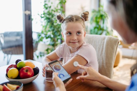 Mädchen mit Diabetes überprüfen den Blutzuckerspiegel zu Hause mit einem kontinuierlichen Blutzuckermessgerät. Mutter eines Mädchens verbindet CGM mit einem Smartphone, um ihren Blutzuckerspiegel in Echtzeit zu überwachen.