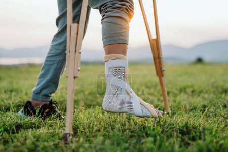 Photo for Close up of broken ankle in cast brace Man standing on healthy leg, while broken leg is in synthetic cast, supported with crutches. - Royalty Free Image
