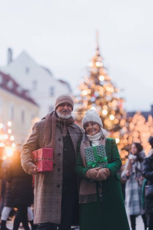 Photo for Happy senior couple enjoying outdoor christmas market in the city, buying gifts. Christmas square with a big Christmas tree in the center. - Royalty Free Image
