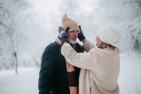 Téléchargez les photos : Élégant couple de personnes âgées marchant dans le parc enneigé, pendant la froide journée enneigée d'hiver. Femme âgée fixant chapeau sur la tête des maris. Paysage hivernal. - en image libre de droit