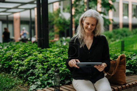 Photo for Portrait of beautiful mature woman in middle age with long gray hair, sitting on the bench in the city. Woman working outdoors, making videocall on tablet. - Royalty Free Image