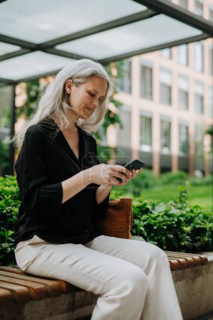 Téléchargez les photos : Portrait de belle femme mûre d'âge moyen avec de longs cheveux gris, assise sur le banc de la ville, tenant un téléphone intelligent. - en image libre de droit