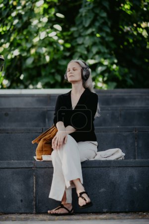 Téléchargez les photos : Portrait d'une belle femme aux cheveux gris, assise sur des escaliers en béton dans la ville, écoutant de la musique à travers des écouteurs sans fil. - en image libre de droit