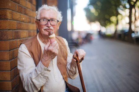 Foto de Retrato de un hombre mayor con bastón, comiendo helado en un caluroso día de verano en la ciudad. Turista mayor en vacaciones de verano en la ciudad. - Imagen libre de derechos