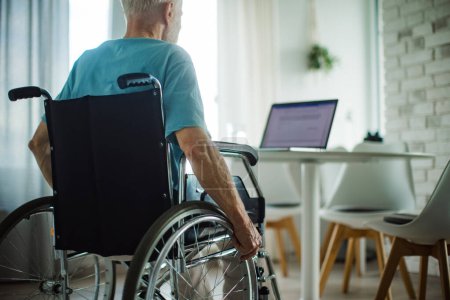 Rear view of senior man in a wheelchair working from home during retirement. Portrait of elderly man using digital technologies, working on a laptop. Concept of seniors and digital skills.