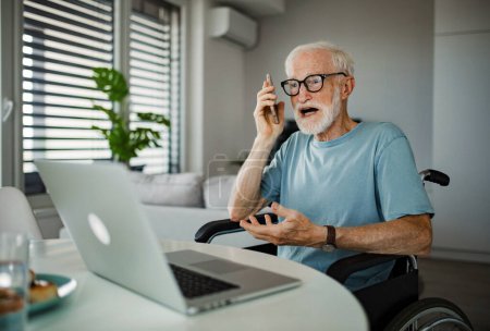 Foto de Hombre mayor en silla de ruedas que trabaja desde casa durante la jubilación. Hombre mayor que usa tecnologías digitales, trabaja en una computadora portátil y hace llamadas telefónicas en casa. Concepto de personas mayores y habilidades digitales. Mayores - Imagen libre de derechos
