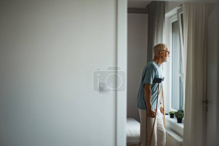 Photo for Sad senior man with crutches spending time alone in his apartment, looking out of window. Concept of loneliness and dependence of retired people. - Royalty Free Image