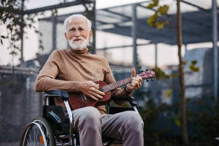 Foto de Hombre mayor sentado en una silla de ruedas al aire libre, tocando el ukelele. Músico retirado tocando la guitarra, recordando los viejos tiempos. Efectos terapéuticos de la música en las personas mayores. - Imagen libre de derechos