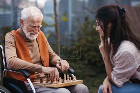Foto de Hombre mayor jugando al ajedrez al aire libre con su hija. Cliente de hogar de ancianos en silla de ruedas que pasa tiempo de calidad con el cuidador, disfrutando en un juego de ajedrez y conversación. - Imagen libre de derechos