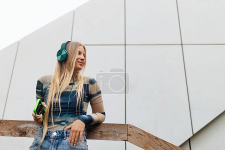 Téléchargez les photos : Portrait d'une étudiante de la génération Z écoutant de la musique via un casque à l'extérieur de la ville. Étudiant passe du temps libre en ligne et seul. Concept de la génération Z comme génération la plus solitaire. - en image libre de droit
