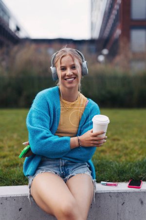 Foto de Retrato de una estudiante de Generación Z escuchando música a través de auriculares inalámbricos al aire libre en la ciudad. Estudiante pasar tiempo libre solo, disfrutando del café en una taza de viaje reutilizable. Concepto de gen Z como - Imagen libre de derechos