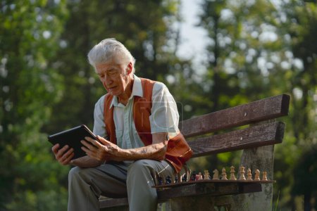 Téléchargez les photos : Homme âgé assis dans le parc sur un banc, regardant des photos sur sa tablette et se sentant nostalgique. Le vieillard solitaire en attente d'un appel vidéo de sa famille. - en image libre de droit
