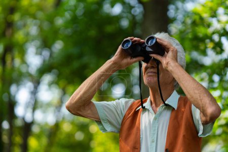 Senior man spending free time outdoors in nature, watching forest animals through binoculars. A retired forest ranger feeling at home in forest, monitoring wildlife and ecological changes.