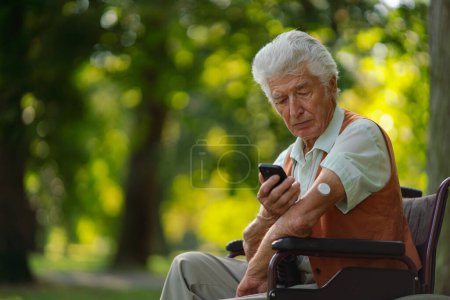 Téléchargez les photos : Homme diabétique aîné en fauteuil roulant vérifiant ses données de glucose sur smartphone. Aîné diabétique utilisant un glucomètre continu pour vérifier le taux de sucre dans le sang. - en image libre de droit