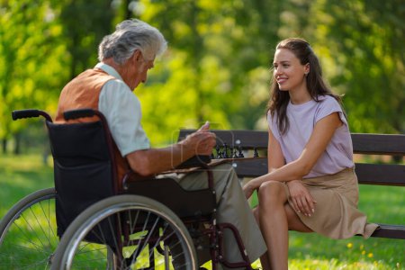 Photo for A young caregiver spending quality time with lonely senior client in the city park. The elderly man in a wheelchair spends time outdoors with his granddaughter, playing chess and chatting. - Royalty Free Image