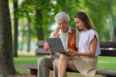Photo for A young caregiver spending quality time with lonely senior client in the city park. The grandfather and granddaughter are looking at photos on a notebook, shopping online together. - Royalty Free Image