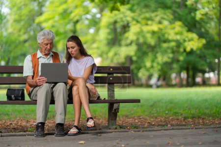 Photo for A young caregiver spending quality time with lonely senior client in the city park. The grandfather and granddaughter are looking at photos on a notebook, shopping online together. - Royalty Free Image