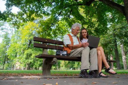 Photo for A young caregiver spending quality time with lonely senior client in the city park. The grandfather and granddaughter are looking at photos on notebook sitting on a bench in a city park. Young - Royalty Free Image