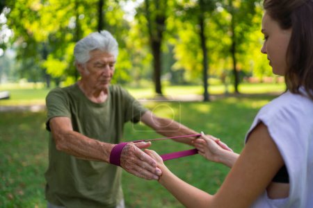 Photo for The senior man exercising in the park, using resistance band. A young caregiver showing older man how to perform the exercise with resistance band correctly. A portrait of a young physiotherapist - Royalty Free Image