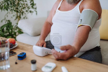 Téléchargez les photos : Homme en surpoids mesurant sa tension artérielle à la maison. Homme ayant une pression artérielle élevée utilisant un tensiomètre à domicile. - en image libre de droit