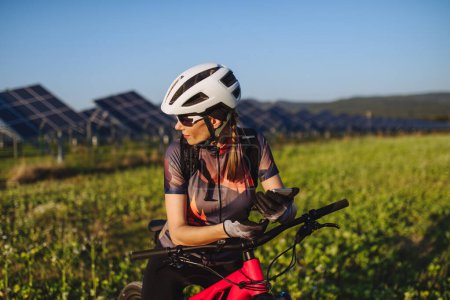 Photo for Portrait of a beautiful cyclist standing in front of solar panels at a solar farm during a summer bike tour in nature. A solar farm as solution for more sustainable energy future. - Royalty Free Image