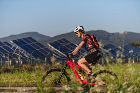 Téléchargez les photos : Vue latérale d'un beau cycliste chevauchant devant des panneaux solaires dans une ferme solaire lors d'une excursion en vélo d'été dans la nature. Une ferme solaire comme solution pour un avenir énergétique plus durable. - en image libre de droit