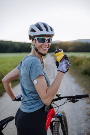 Téléchargez les photos : Cycliste diabétique avec un glucomètre continu sur son bras boire de l'eau pendant sa tournée à vélo pour gérer son diabète tout en faisant de l'exercice. Concept d'exercice et de diabète. - en image libre de droit