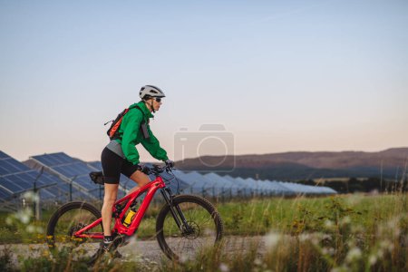 Foto de Vista lateral de un hermoso ciclista cabalgando frente a paneles solares en una granja solar durante un recorrido en bicicleta de verano en la naturaleza. Una granja solar como solución para un futuro energético más sostenible. - Imagen libre de derechos